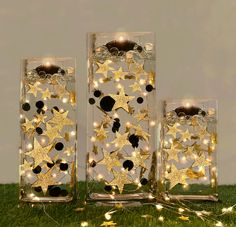 three clear glass vases with gold stars and lights in them on green grass next to wall