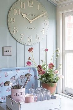 a clock on the wall above a kitchen counter with flowers and utensils in front of it