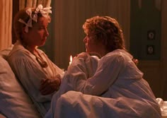 two women in white robes sitting on a bed next to each other and one is holding a lit candle