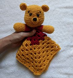 a hand holding a crocheted teddy bear on top of a white blanket with a red ribbon