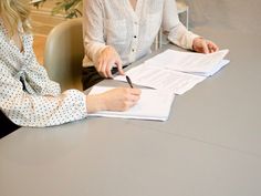 two women sitting at a table with papers in front of them and one holding a pen