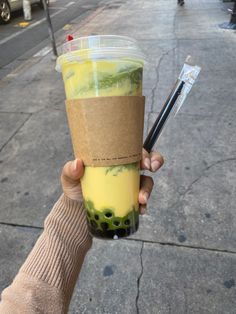 a person holding up a drink in their hand on the sidewalk with other people walking down the street behind them