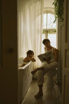 two people sitting on a toilet in a bathroom reading a newspaper next to each other