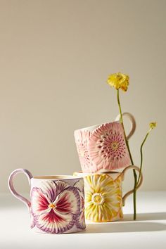 three coffee mugs with flowers in them on a white surface, one is pink and the other is yellow