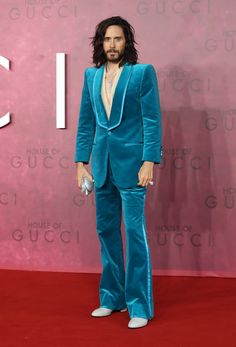 a man with long hair wearing a blue suit and white shoes standing on a red carpet