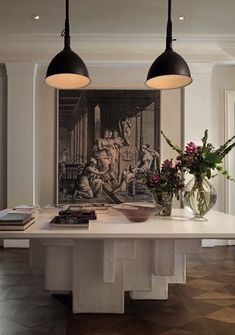 two lamps hanging over a white table with flowers in vases on the top and paintings behind it