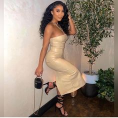 a woman in a tan dress leaning against a wall next to a potted plant