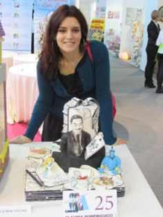 a woman standing next to a table with books on it and a cake in the shape of a man's face