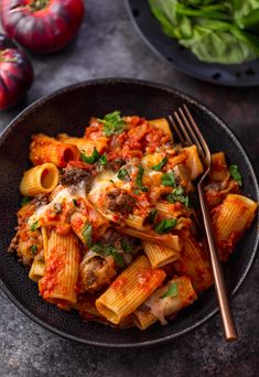 a black plate topped with pasta and meat covered in tomato sauce next to some spinach