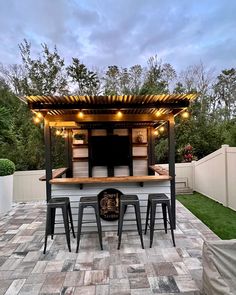 an outdoor bar with stools and lights on the outside wall, next to a fence