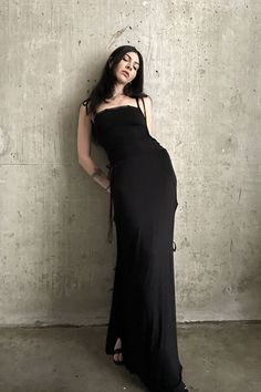 a woman in a long black dress leaning against a wall with her hands on her hips