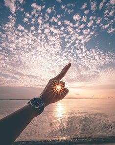 a person's hand pointing at the sun in the sky over water with clouds