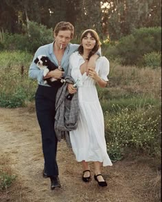 a man and woman walking down a dirt road with a cat in their arms,