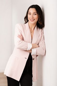 a woman in a pink jacket leaning against a wall with her arms crossed and looking at the camera