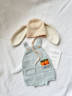 a knitted bunny hat and overalls are laying on a white sheet with a card