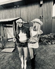 two women in cowboy hats and shorts standing next to each other on the grass near a barn