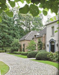 a large house with lots of trees and bushes in front of it on a cobblestone driveway