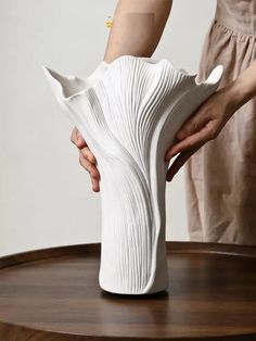 a white vase being held by two hands on top of a wooden table with a woman in the background