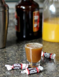 a shot glass filled with liquid next to two candy bars on a counter top and bottles of orange juice in the background