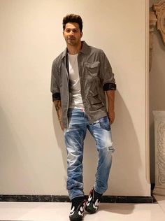 a man standing in front of a white wall wearing ripped jeans and a gray jacket