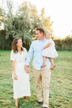 a man and woman walking through a field with their baby girl in her arms as the sun sets behind them