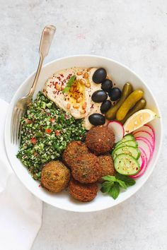 a white bowl filled with meatballs, vegetables and pickles next to a fork