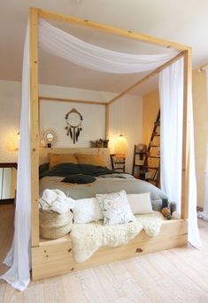 a canopy bed with white drapes and pillows on the bottom, in a bedroom