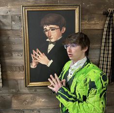 a young man wearing glasses standing in front of a painting with his hands folded up