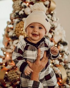 a baby wearing a plaid shirt and hat standing in front of a christmas tree