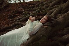 a woman laying on top of a tree trunk in the forest wearing a white dress