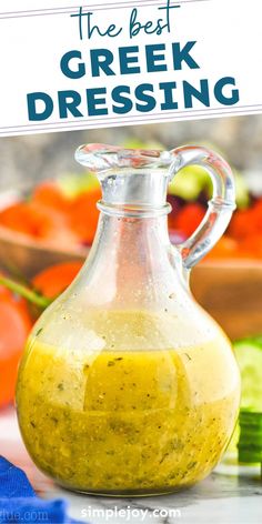 the best greek dressing recipe in a glass pitcher on a table with vegetables behind it