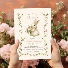 a person holding up a baby shower card in front of pink flowers and greenery