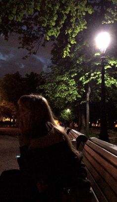 a woman sitting on a bench at night with the lights turned on and trees in the background