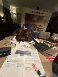 a woman sitting at a desk covered in papers and pens, with her head on top of a pile of paperwork