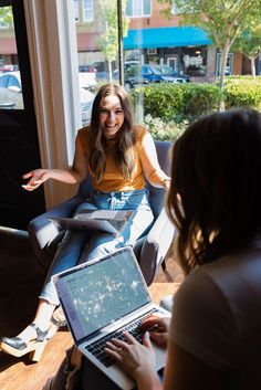 two women sitting in front of a window with laptops on their laps and talking