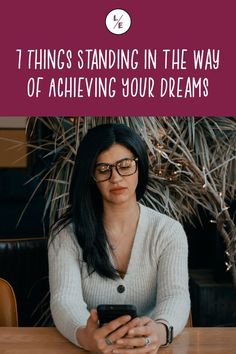 a woman sitting at a table looking at her cell phone with the text, 7 things standing in the way of achieving your dreams