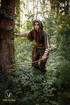 a woman dressed in medieval clothing holding a bow and arrow while standing next to a tree