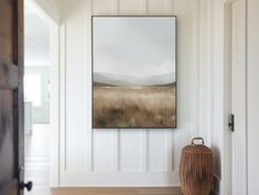 a painting hanging on the wall above a basket in a room with white walls and wood flooring