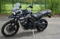 a black motorcycle parked on top of a parking lot next to a green fence and trees