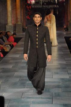 a man is walking down the runway in a black suit and long pants with gold buttons