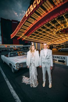 two people standing next to each other in front of a white car at night time