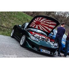 two men standing next to a green sports car with its hood open and it's engine in the trunk