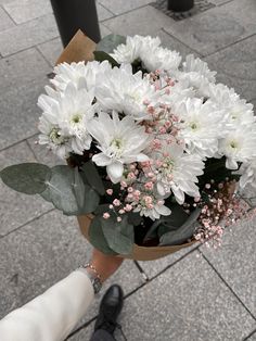 a person holding a bouquet of white flowers and greenery in their hand on the sidewalk