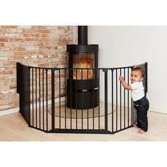 a baby standing in front of a black gate next to a brick wall and fireplace