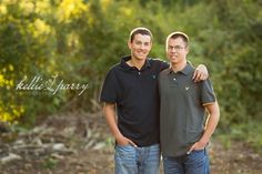 two men standing next to each other in front of trees and bushes with their arms around each other