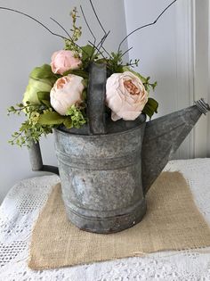 a metal watering can with flowers in it on a doily covered tablecloth next to a window