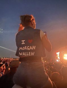 a man with his back to the camera at an outdoor concert, wearing a vest that says i love morgan wallen