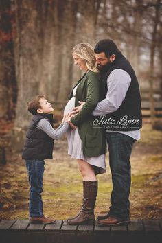 a pregnant woman and her husband hold their son's hand as they stand on a bridge in the woods