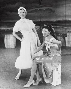 two women are posing for the camera in front of some barrels and barrells, one is wearing a white dress with polka dots on it