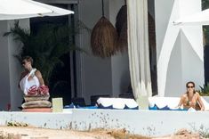 two women sitting under umbrellas on the beach in front of a building with palm trees